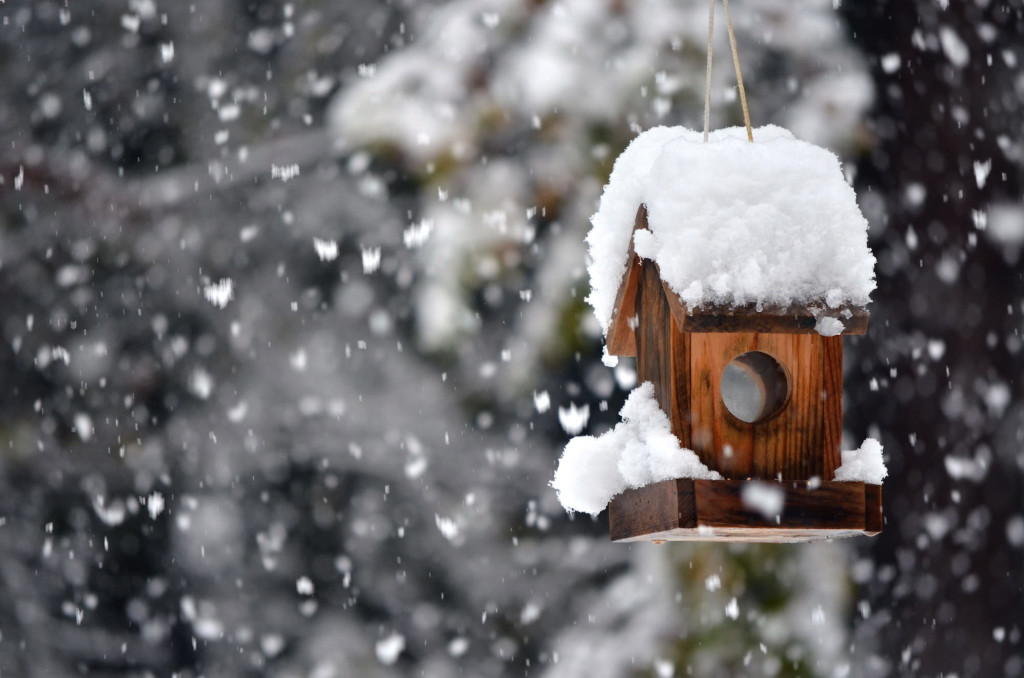 A snow covered bird house in winter with snowflakes falling down.